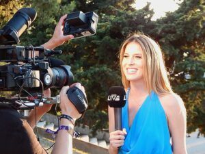 Young reporter dressed in blue and standing in front of several cameras.