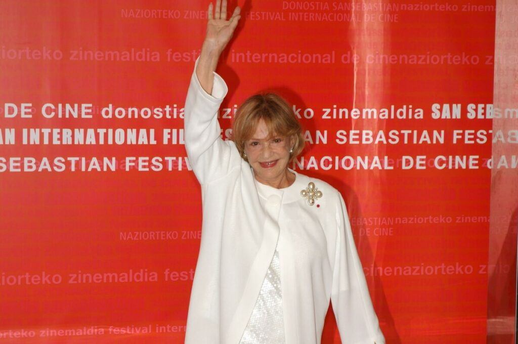 Old actress dressed in white and waving her right hand.